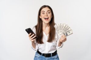 a young, happy woman holding a mobile phone and dollar bills in her hands